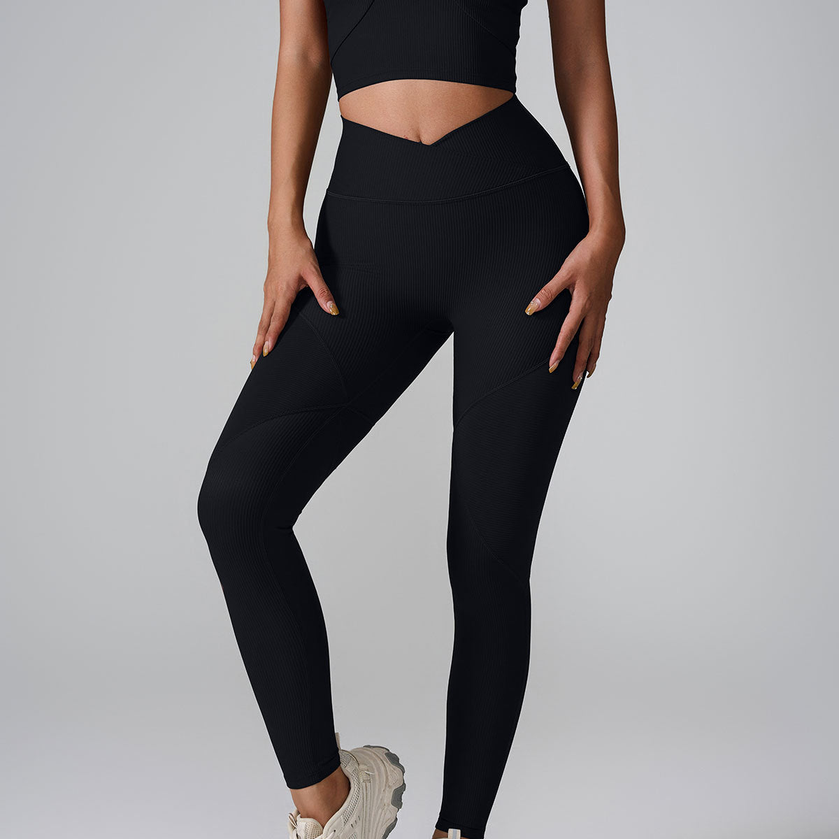 A model wearing black ribbed leggings with V-shaped waistband and matching crop top against gray backdrop