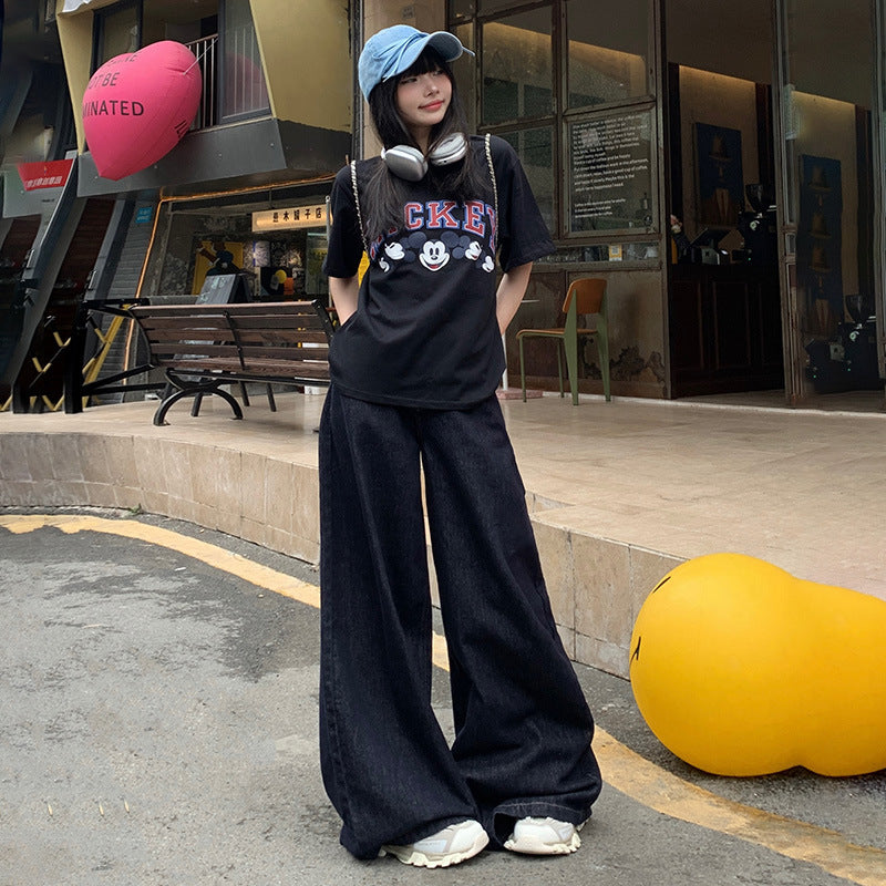 A woman is wearing wide-leg dark denim jeans paired with Mickey Mouse graphic t-shirt, baseball cap and white sneakers, casual streetwear look outside shop