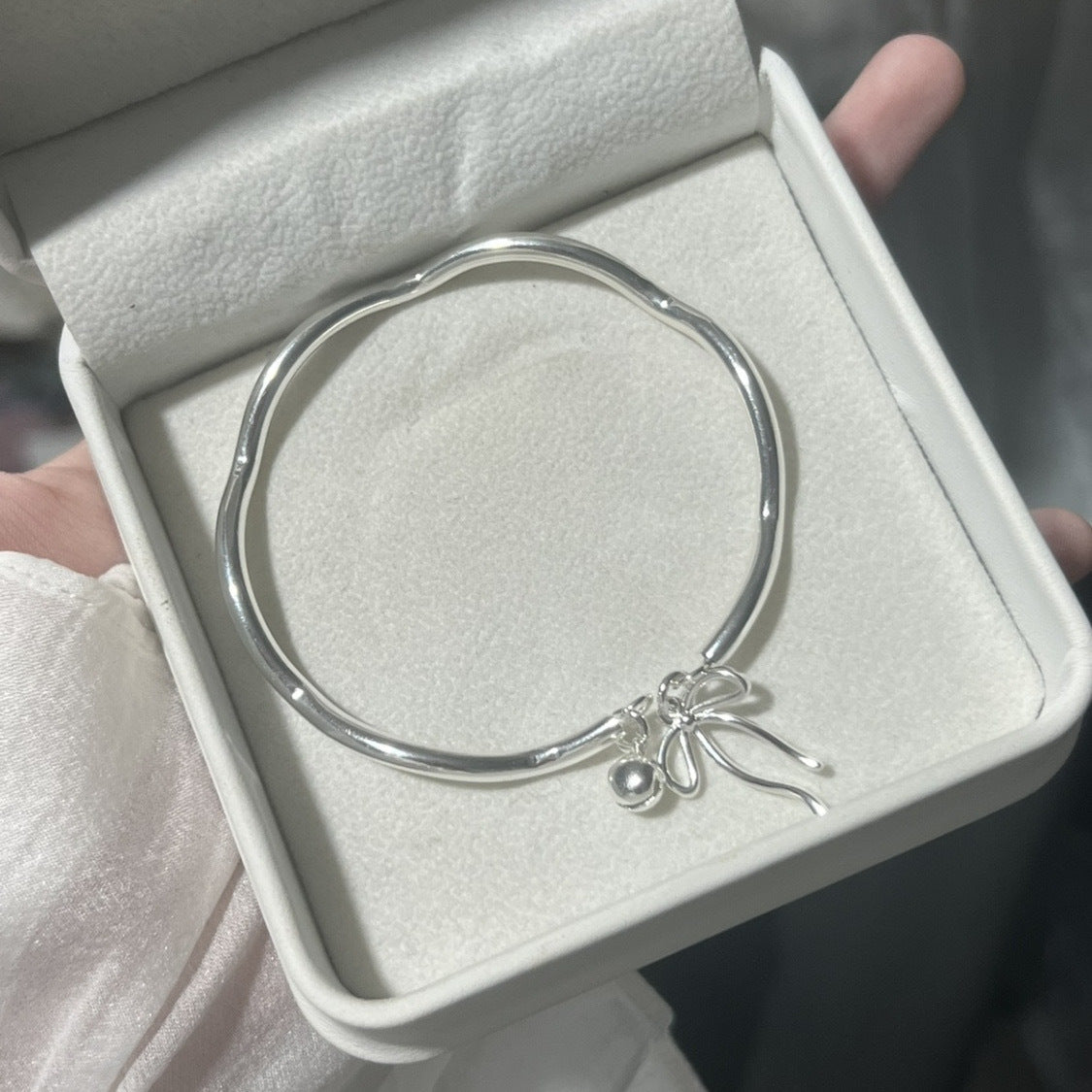 A silver bangle bracelet with bow and bell charms displayed in a white jewelry box with padded interior