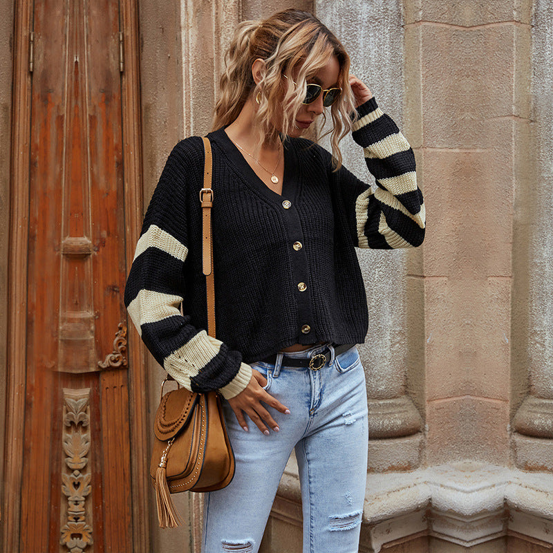 A woman wearing a black cardigan with cream striped sleeves stands against a stone building backdrop, paired with light denim jeans and tan crossbody bag