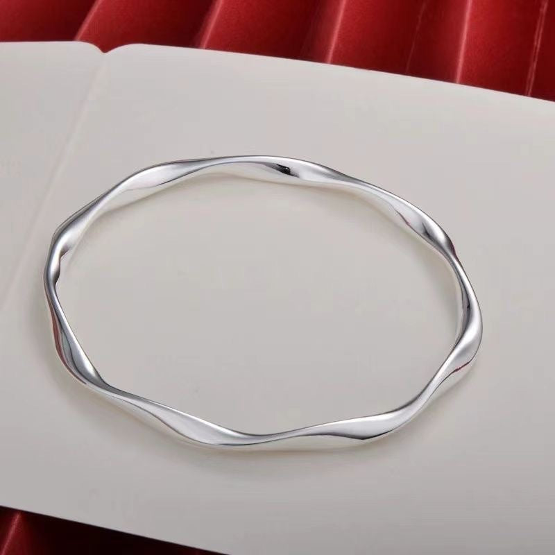 A silver twisted bangle bracelet displayed on a white page, featuring a minimalist octagonal design with gentle curves and high-polish finish
