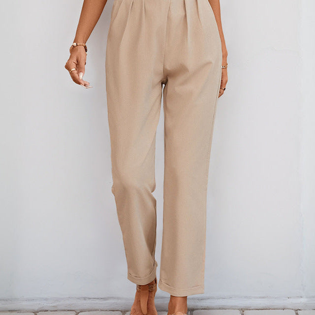 A woman wearing high-waisted beige pleated trousers with a white top and tan sandals, posing against a white wall.