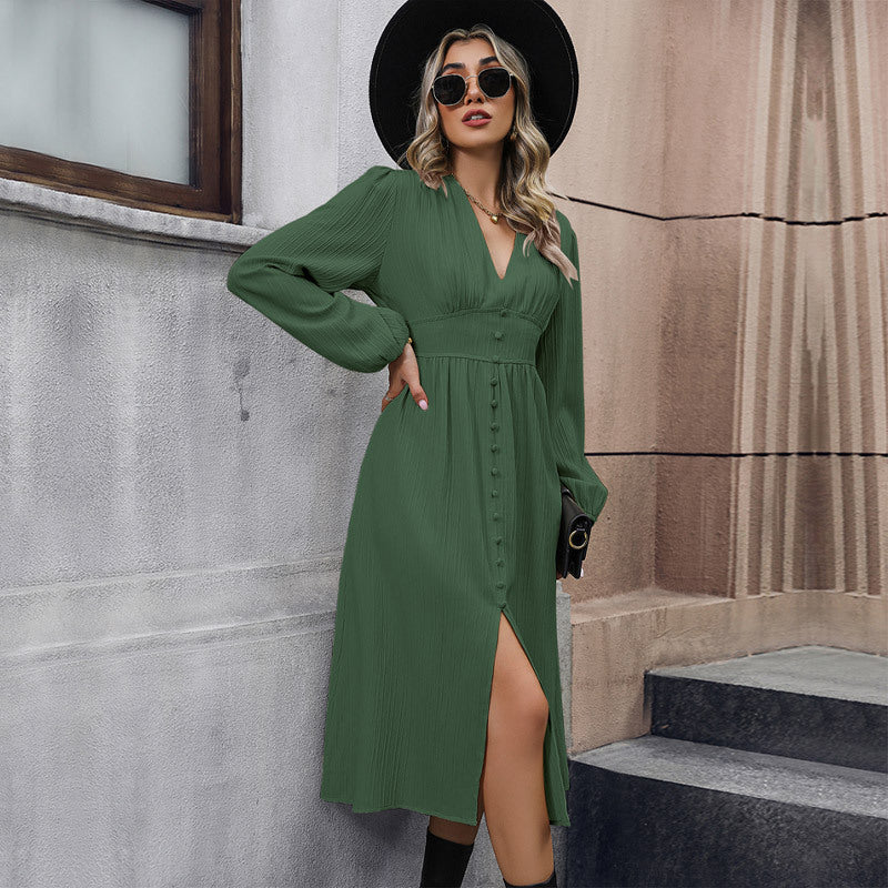 Woman in olive green midi dress, black hat, and sunglasses, leaning against concrete wall