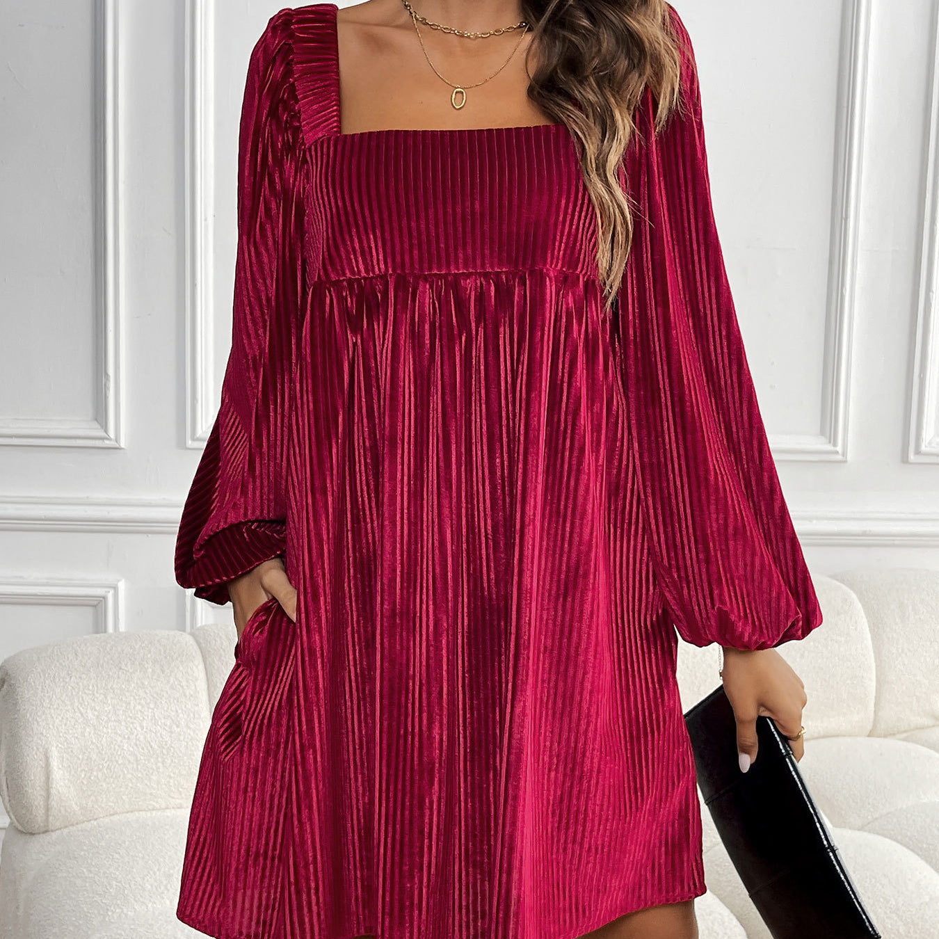 A woman wearing a burgundy ribbed velvet babydoll dress with balloon sleeves stands against white paneling, holding a black clutch