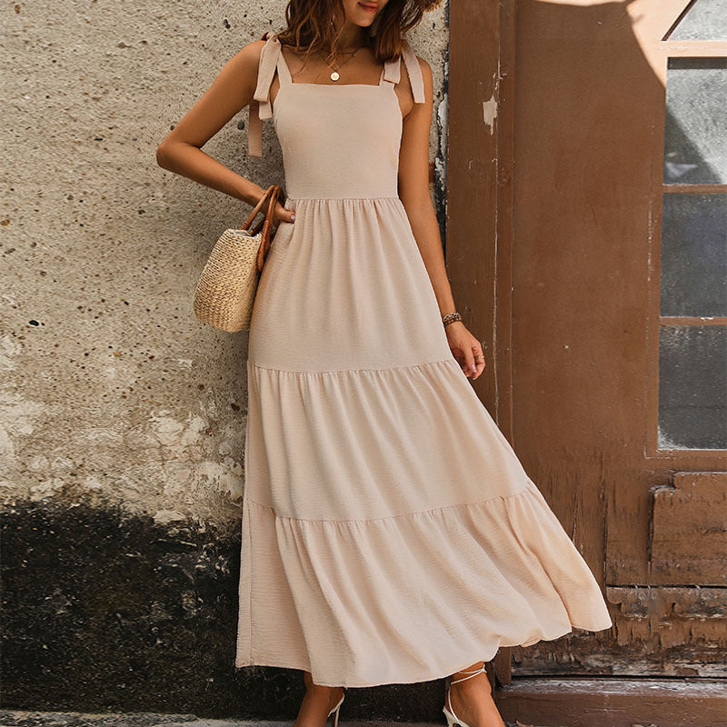 A woman is posing against rustic wall in beige tiered maxi dress with tie shoulder straps, styled with straw basket bag and neutral accessories