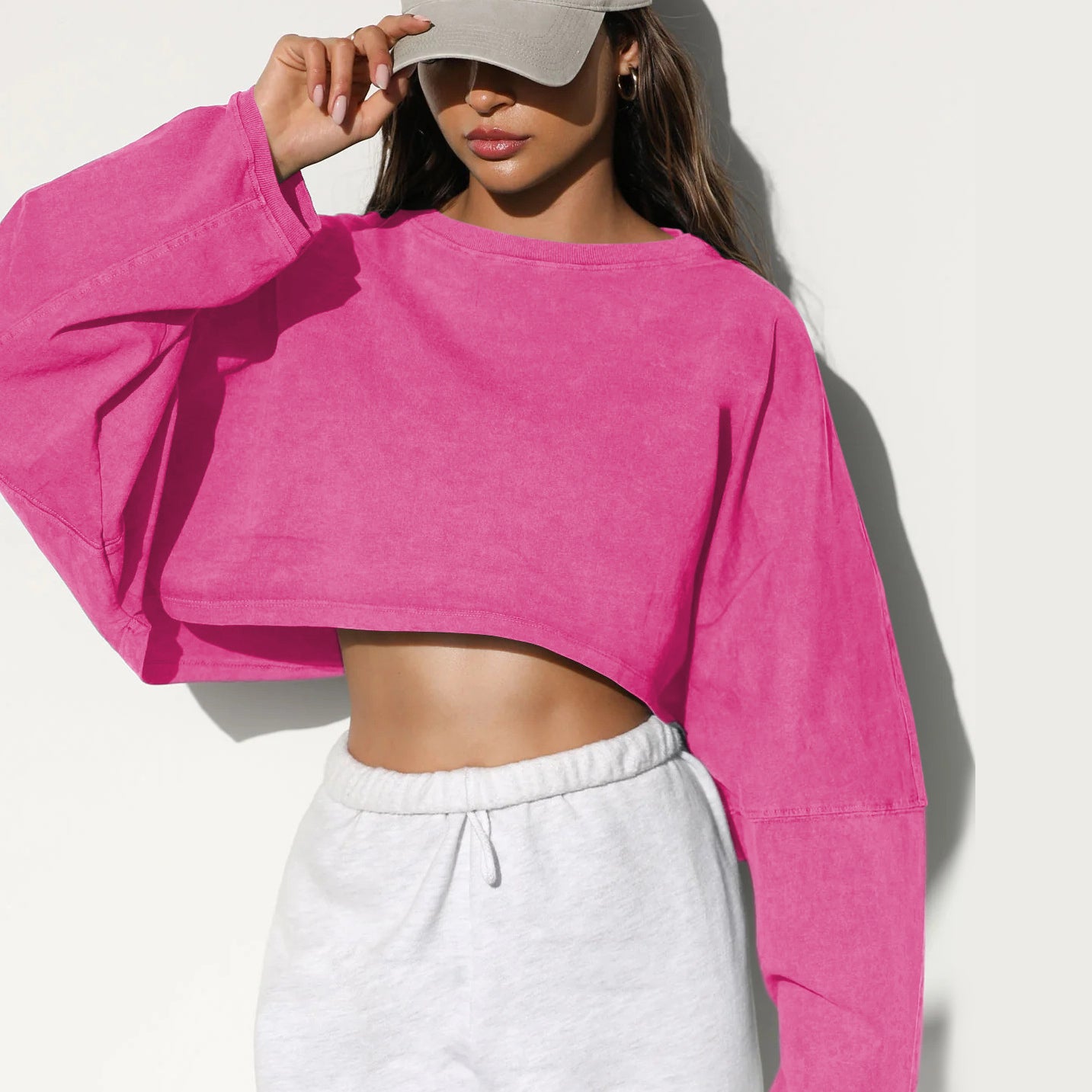 A woman wearing a bright pink cropped long sleeve top and white sweatpants touches her beige baseball cap against a white background