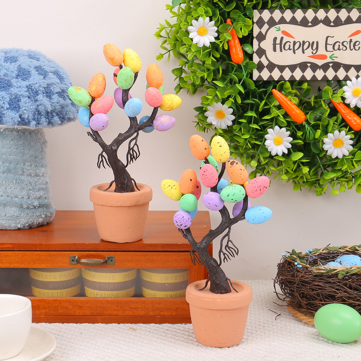 Two Easter egg trees in terracotta pots with multicolored speckled eggs, displayed on a wooden shelf with a green wreath and festive decorations in the background.