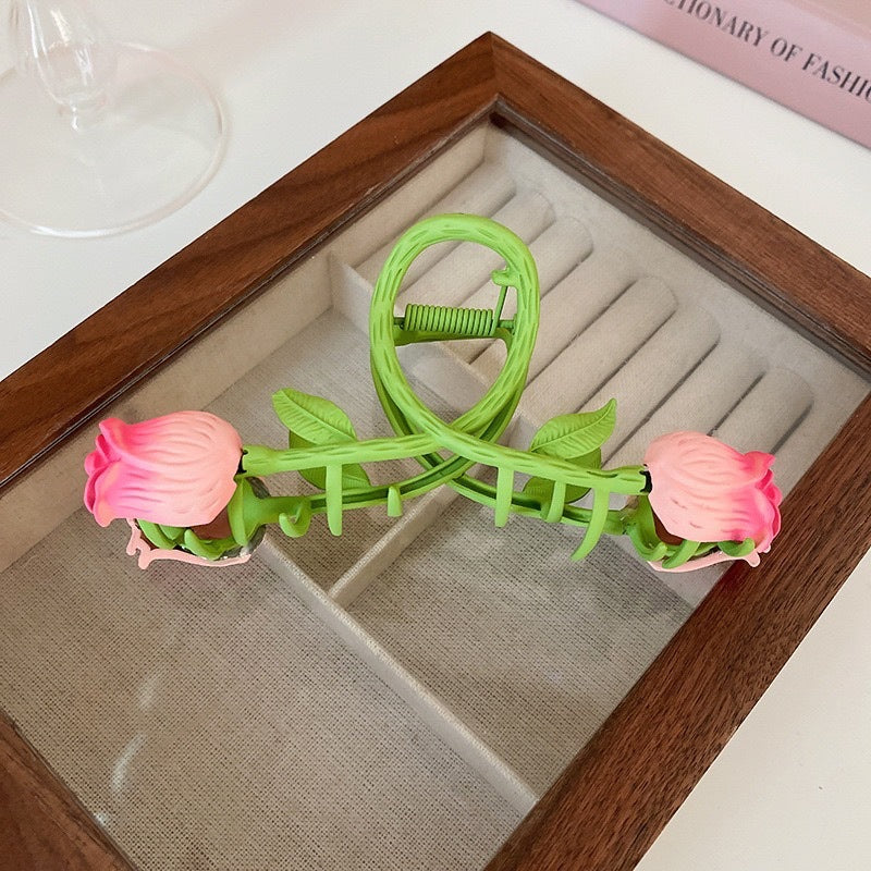 A unique green hair claw designed like a rose vine, featuring two pink rosebuds at each end, displayed on a jewelry tray.