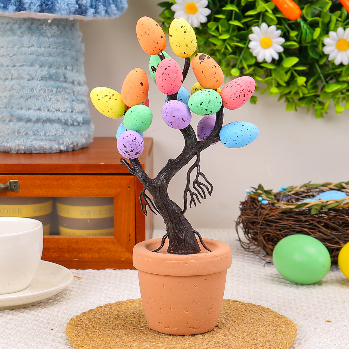 A small tabletop Easter tree featuring colorful speckled eggs on dark branches, placed in a rustic terracotta pot with spring-inspired decorations around it.