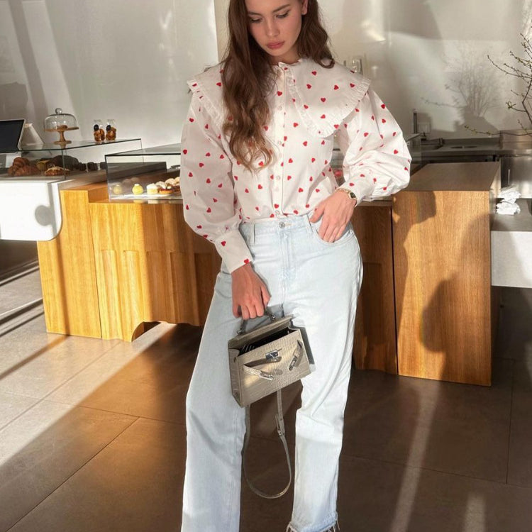 A woman wearing a white heart-print blouse with ruffle collar stands in a café, holding a gray handbag with light denim jeans and cream shoes