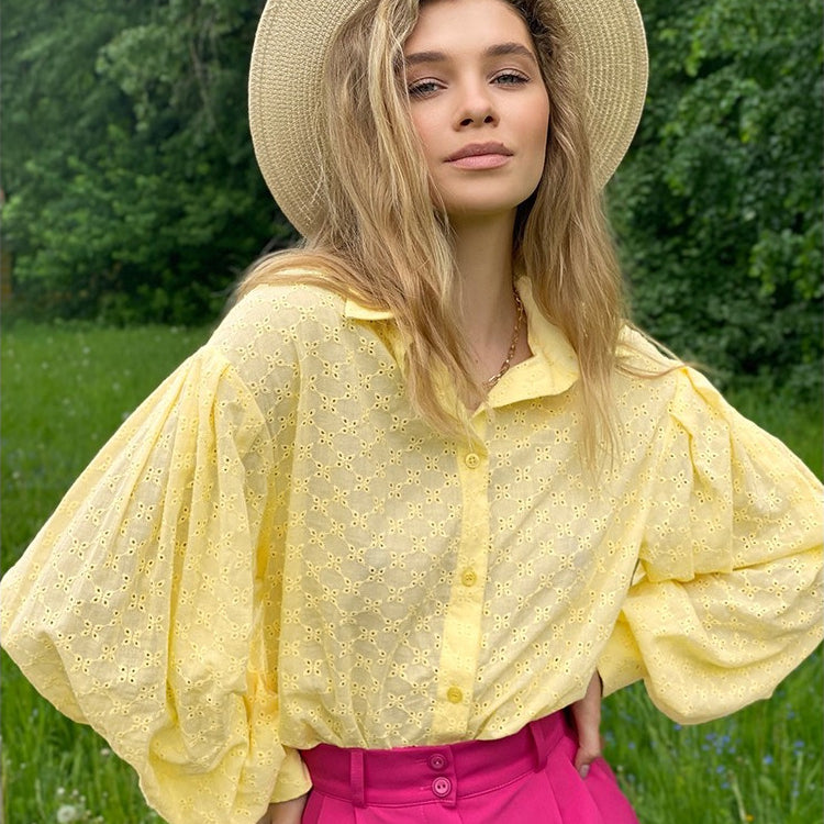 A woman wearing a yellow eyelet button-down shirt with balloon sleeves and bright pink pants posing outdoors in a grassy field wearing a straw hat