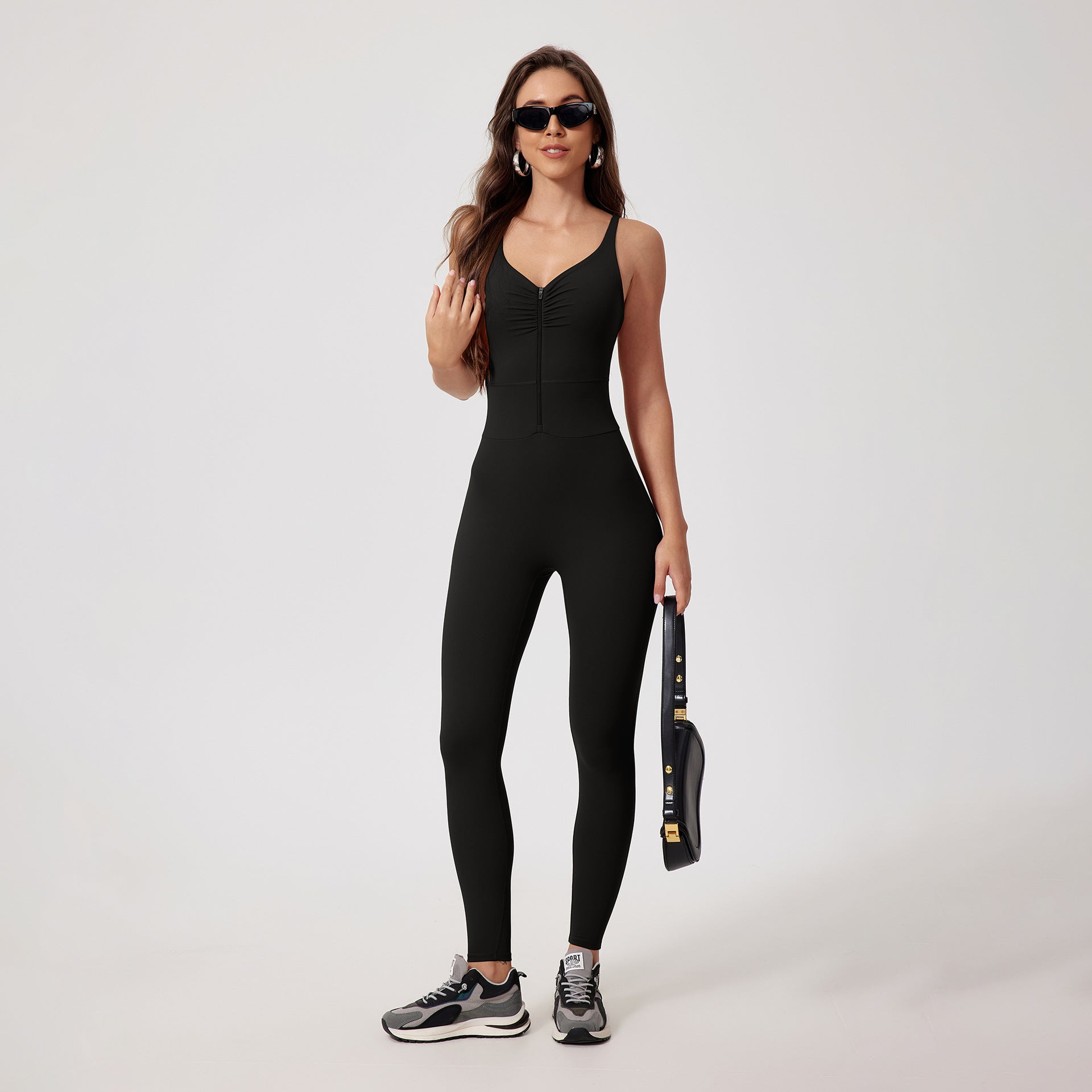 A woman wearing black athletic jumpsuit with ruched neckline, holding small bag and wearing sunglasses on white backdrop