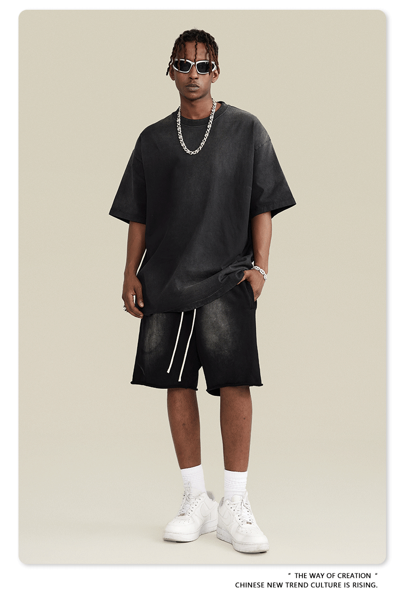 Model with short locs wearing black t-shirt and shorts set with white sunglasses, silver chain necklace and white sneakers against cream background