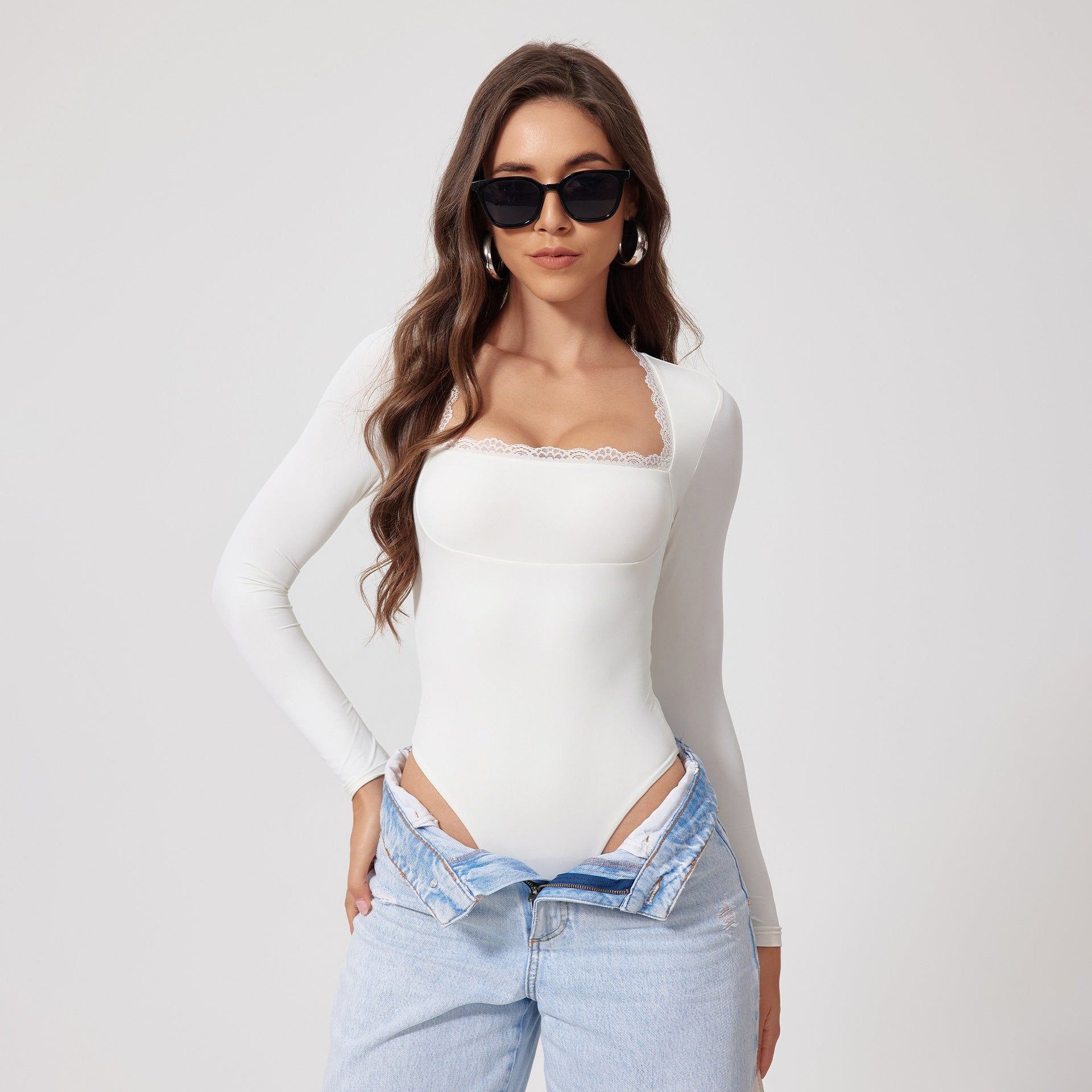 Model in white long-sleeve bodysuit with lace trim neckline, styled with light jeans, sunglasses, and wavy brown hair