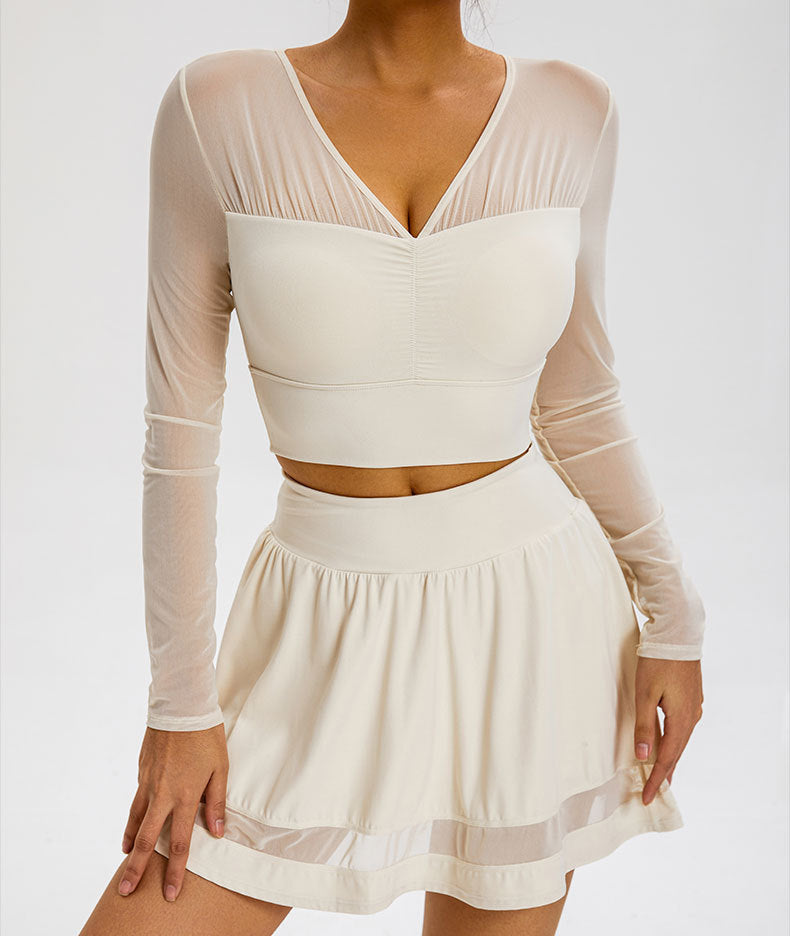 A model is wearing a cream mesh-sleeve crop top with V-neckline and matching flared mini skirt with sheer panel detail, posed against a light background