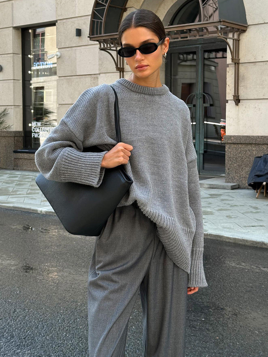 A model is wearing a gray oversized sweater with wide-leg pants while standing on city street. Black handbag and sunglasses complete the monochrome look