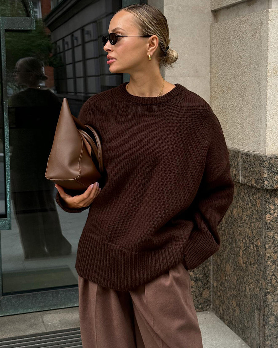 A model is posing in a chocolate brown knit sweater with matching trousers against stone building. Hair in sleek bun with small sunglasses