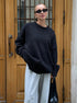 A model is standing before wooden doors wearing a black oversized sweater with light gray pants. Black sunglasses and handbag visible