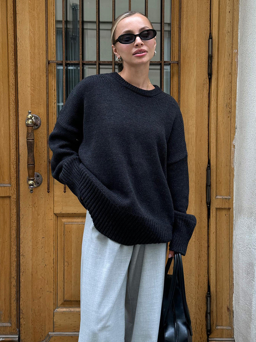 A model is standing before wooden doors wearing a black oversized sweater with light gray pants. Black sunglasses and handbag visible