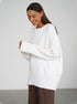 A model is smiling while adjusting white oversized sweater cuffs paired with brown pants against white background. Side profile pose