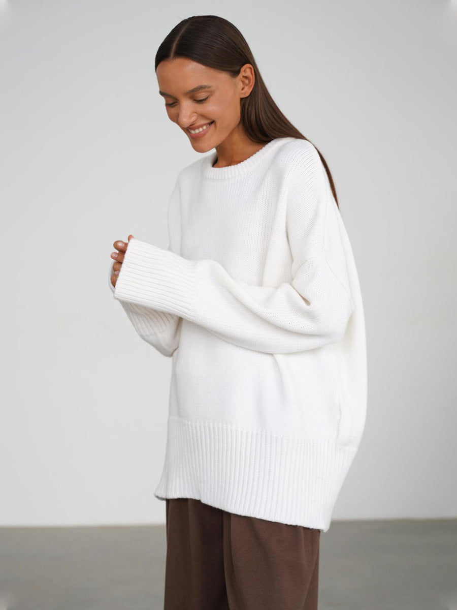 A model is smiling while adjusting white oversized sweater cuffs paired with brown pants against white background. Side profile pose