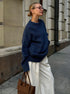 A model is looking upward wearing navy blue knit sweater with white wide-leg trousers on city street. Brown tote bag and glasses visible