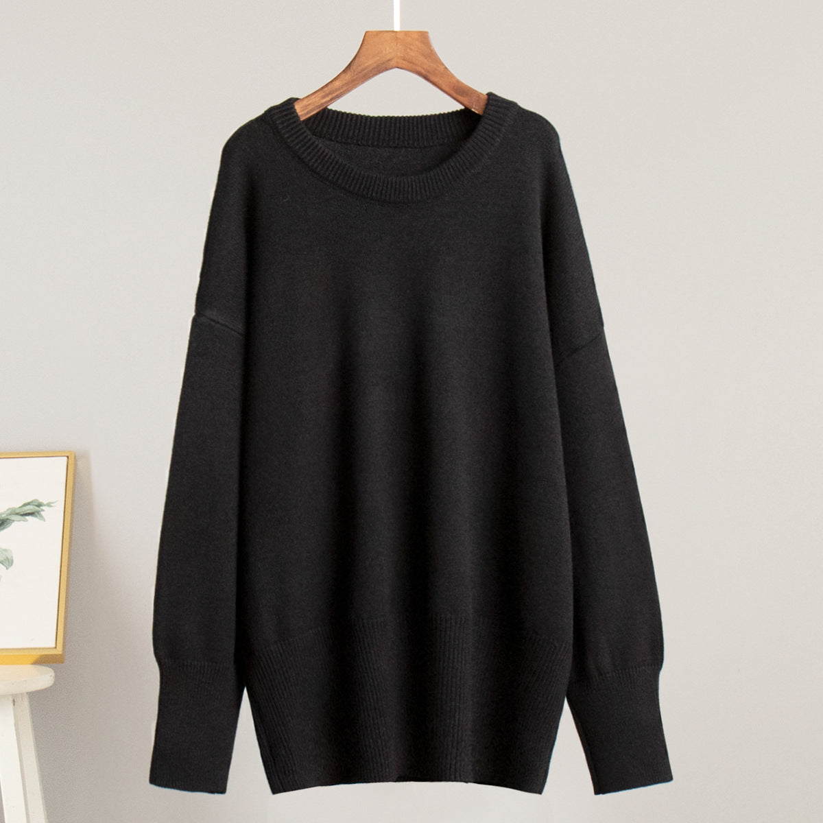 A black oversized sweater displayed on wooden hanger against light background. Classic crew neck with wide ribbed cuffs and hem