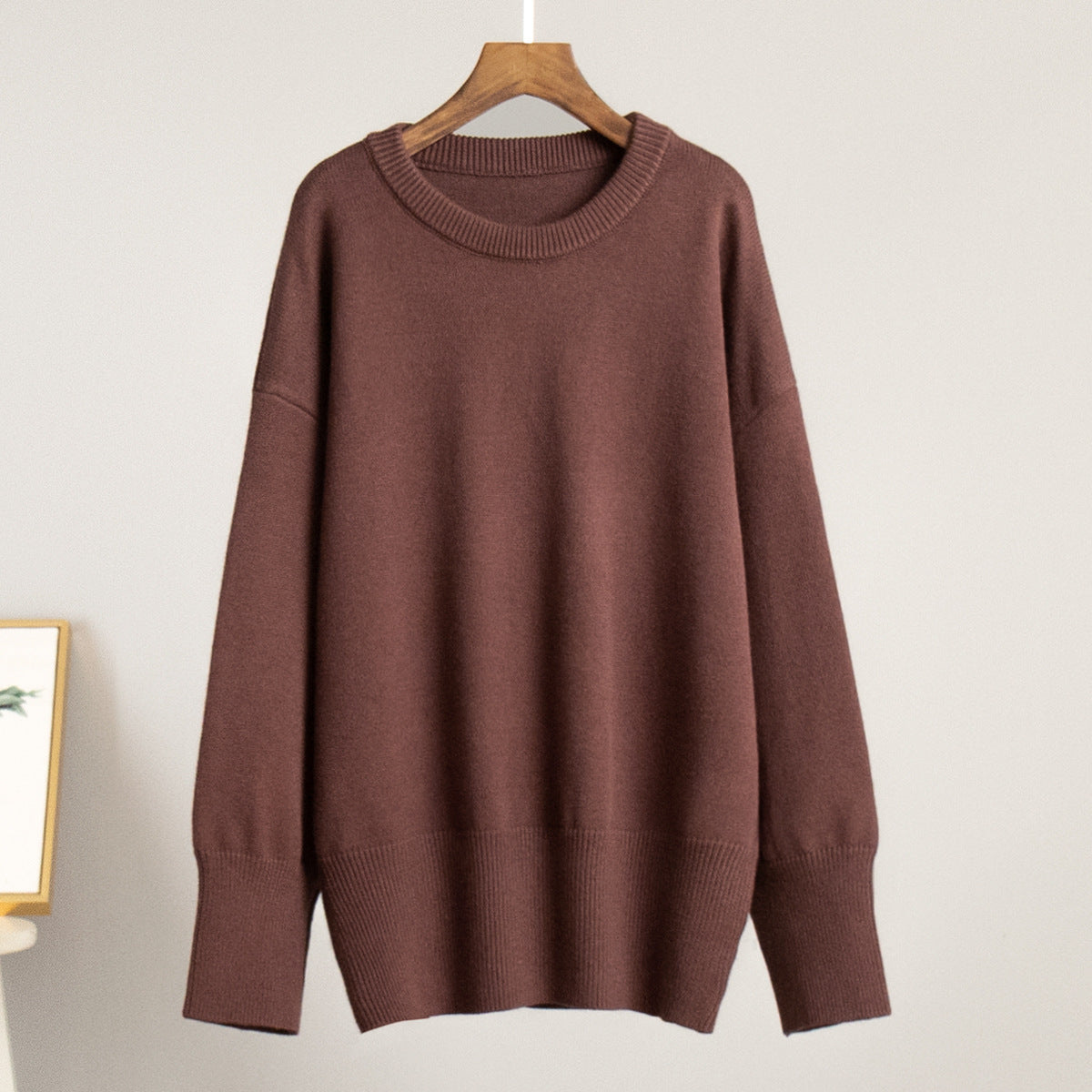 A chocolate brown oversized sweater displayed on wooden hanger against white wall. Drop-shoulder design with ribbed neckline and cuffs