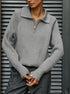 A woman is wearing a gray ribbed zip-collar sweater with gray trousers, standing in front of a dark louvered door