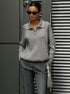 A woman is wearing a gray ribbed zip-collar sweater and dark sunglasses, standing against a dark modern background with a gray handbag