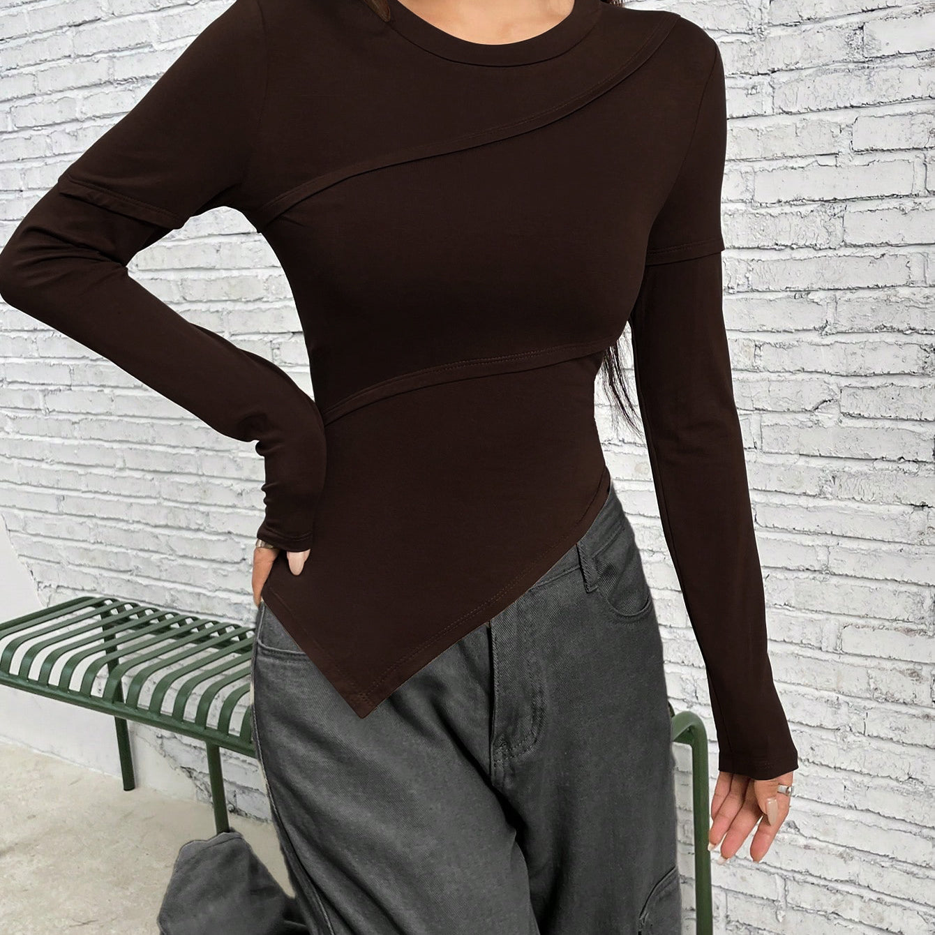 Dark brown asymmetrical long-sleeve top with angular design, styled with gray cargo pants against white brick wall