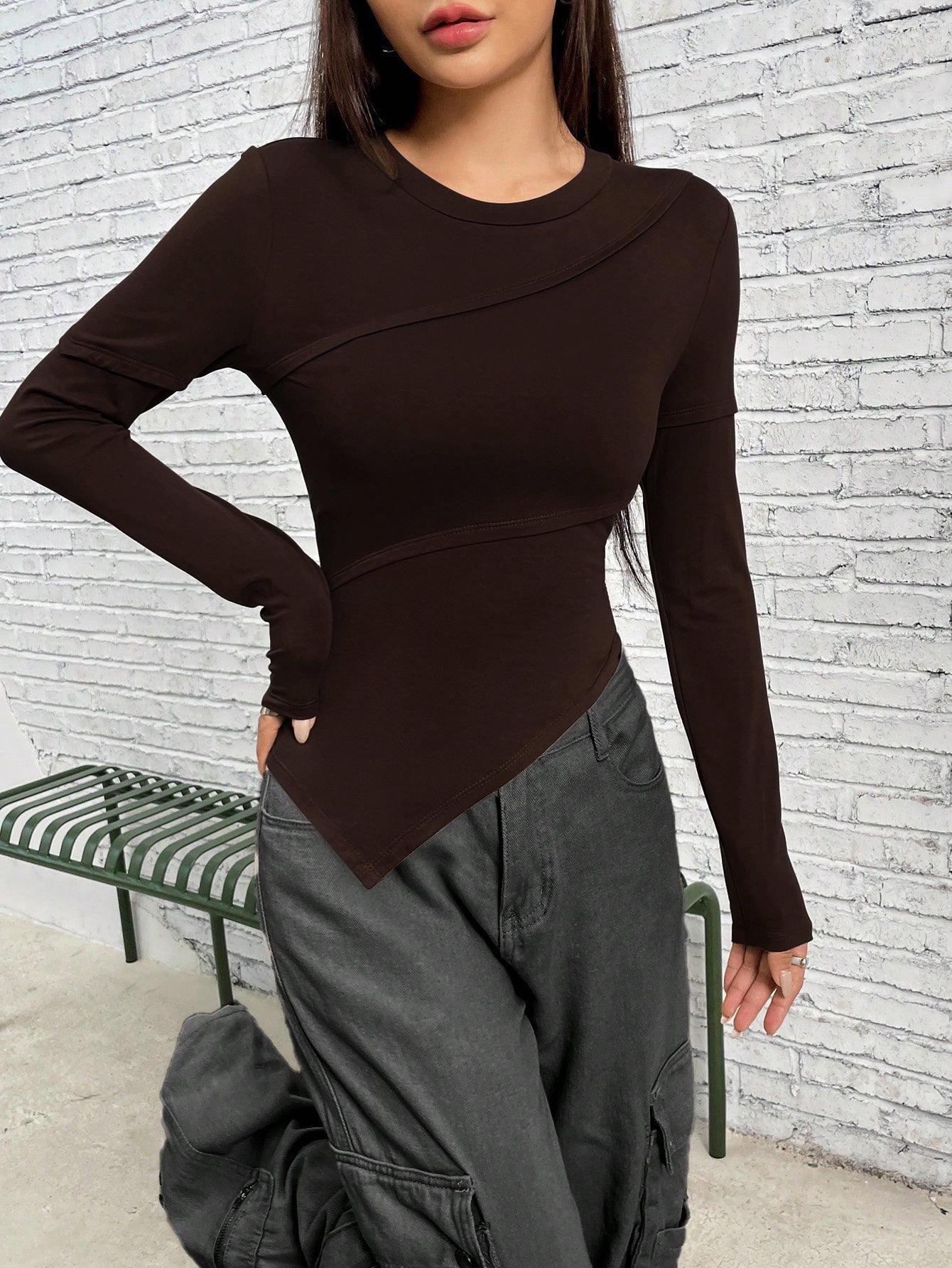 Dark brown asymmetrical long-sleeve top with angular design, styled with gray cargo pants against white brick wall