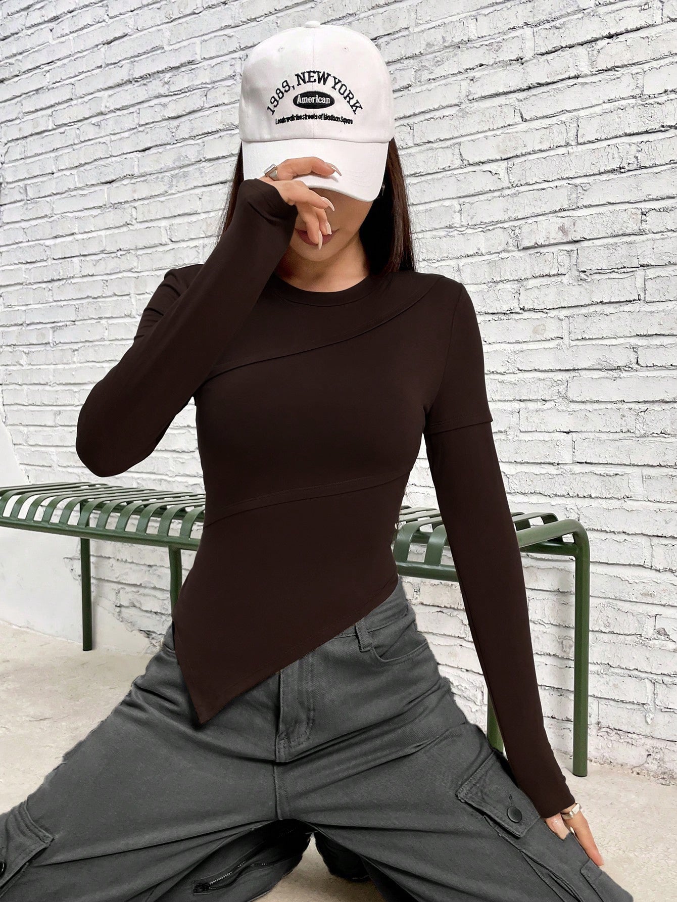 Model in white New York cap, brown asymmetrical top, and gray cargo pants, hand partially covering face