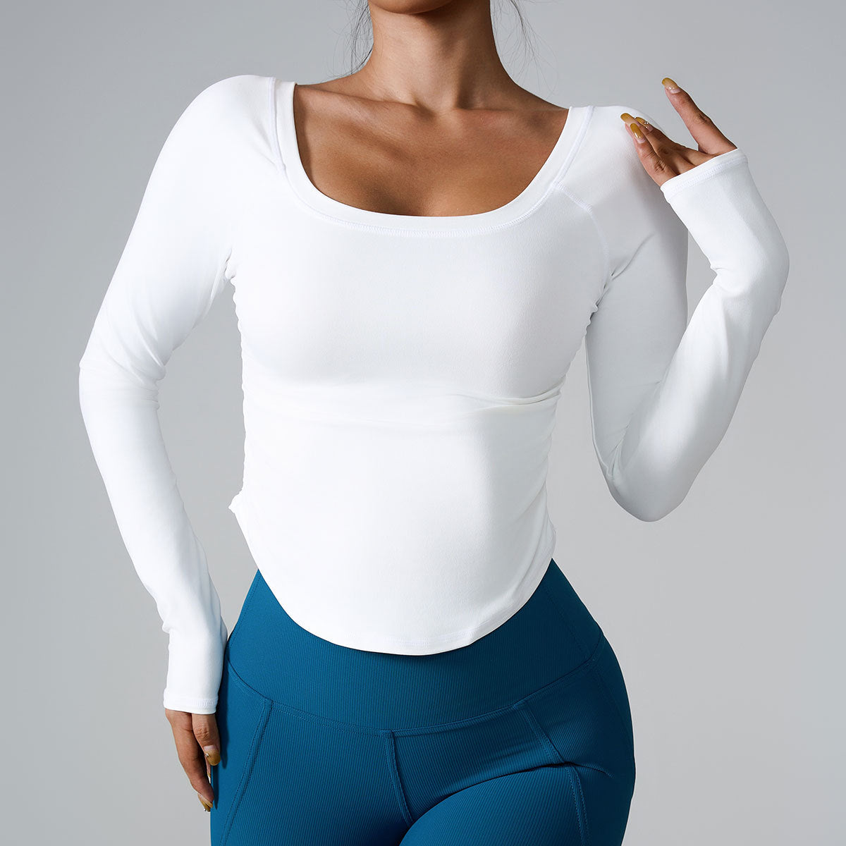 A model wearing a white long-sleeve fitted top with scoop neckline, paired with blue high-waisted athletic leggings, posing against gray backdrop