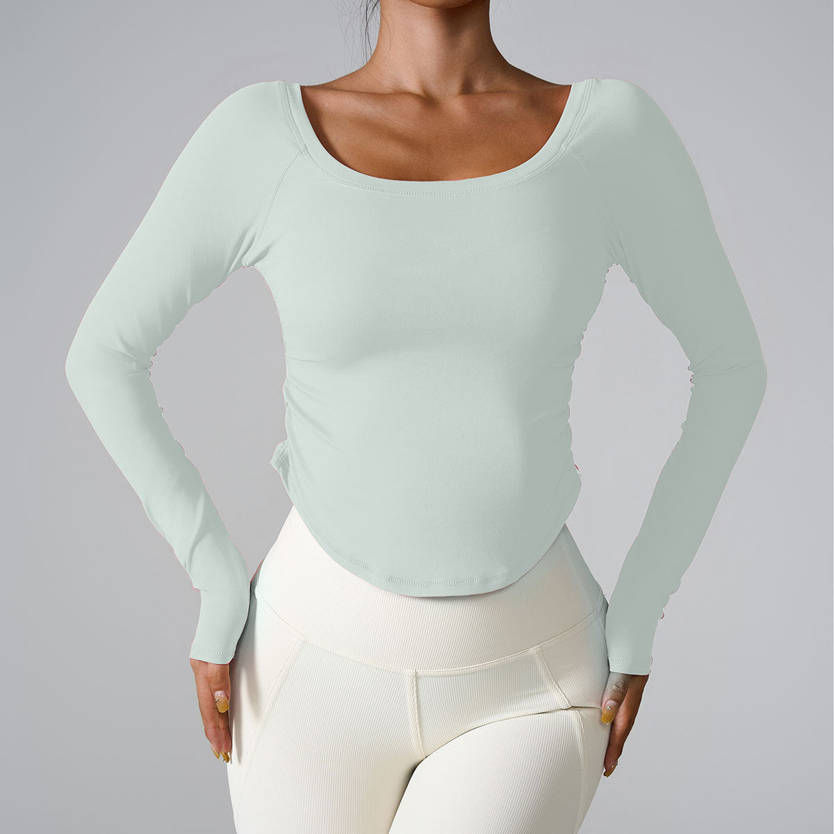 A model wearing a mint green long-sleeve fitted top with scoop neckline, styled with white high-waisted leggings, photographed on gray backdrop