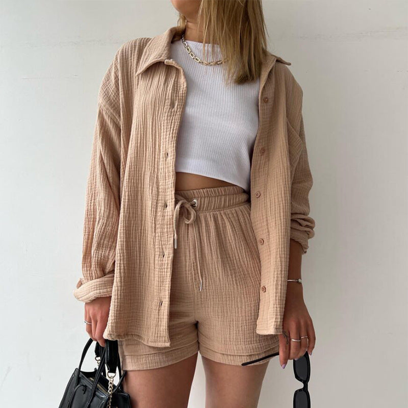 A model is wearing a beige three-piece loungewear set with a white crop top, holding black sunglasses and a black handbag