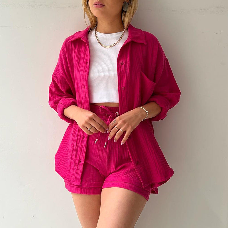 A model is wearing a fuchsia pink three-piece loungewear set with a white crop top, accessorized with gold jewelry and multiple rings