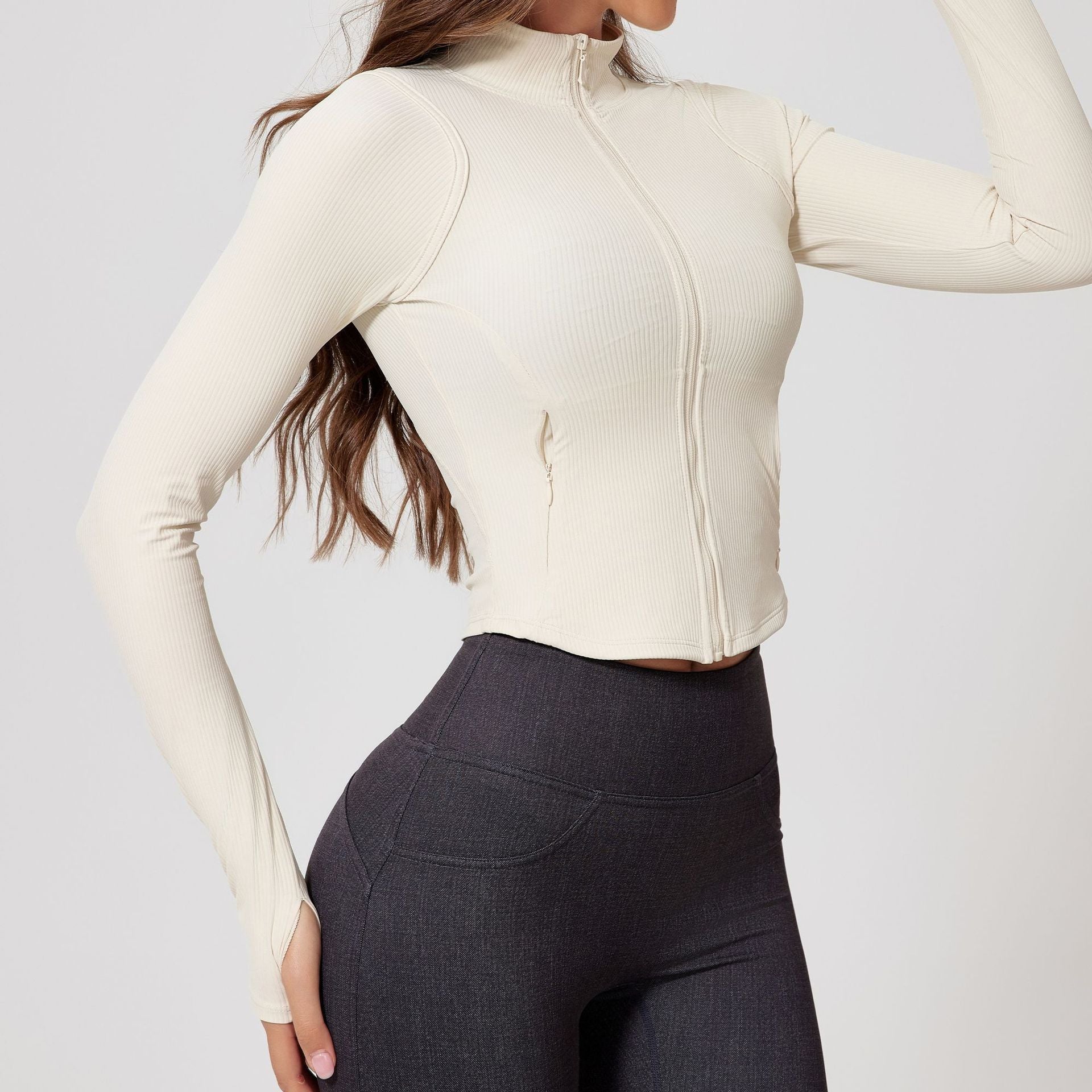 Side view of a woman wearing an ivory ribbed zip-up cropped jacket with long sleeves and thumb holes, paired with dark gray leggings