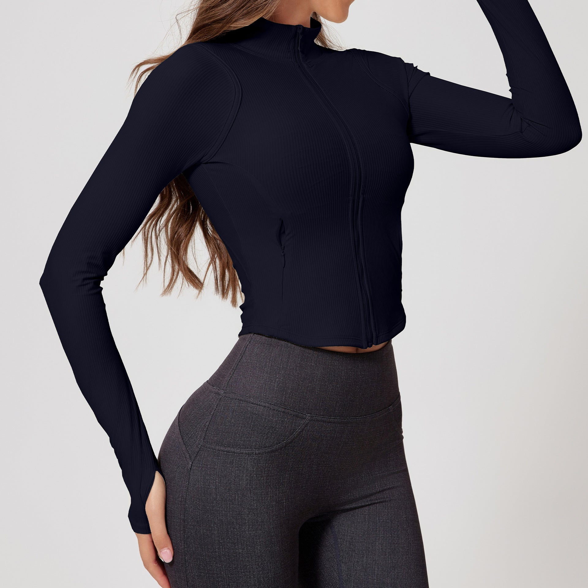 Side view of a woman in a black ribbed zip-up cropped jacket with long sleeves and thumb holes, paired with dark leggings