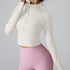 A woman wearing a cream ribbed half-zip crop top with mock neck and long sleeves paired with pink high-waisted leggings against a gray background