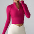 A woman wearing a raspberry pink ribbed half-zip crop top with stand collar, adjusting the zipper, paired with cream leggings against gray backdrop