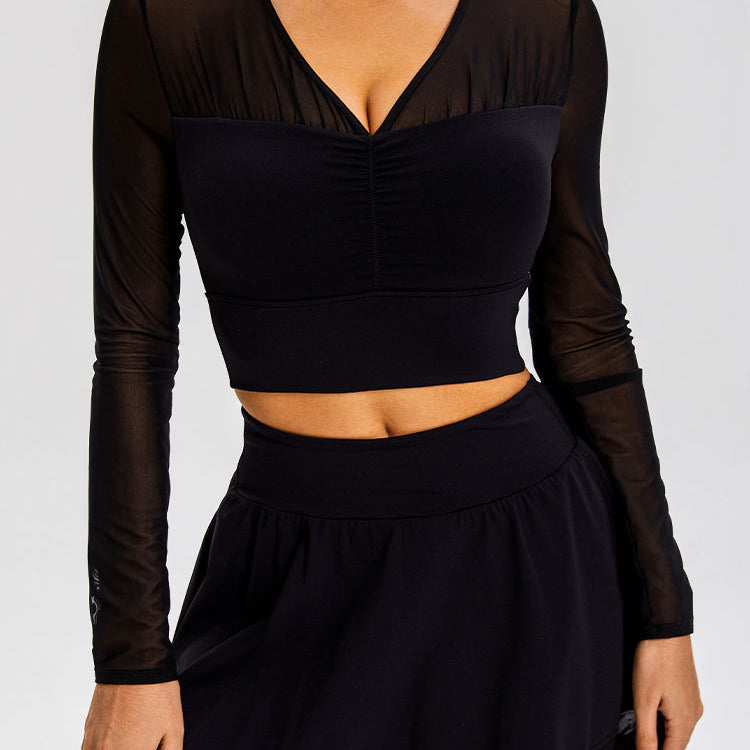 A model is wearing a black mesh-sleeve crop top with V-neckline and gathered center detail, paired with a black mini skirt, against a gray backdrop