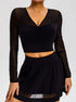A model is wearing a black mesh-sleeve crop top with V-neckline and gathered center detail, paired with a black mini skirt, against a gray backdrop