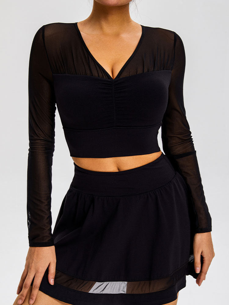 A model is wearing a black mesh-sleeve crop top with V-neckline and gathered center detail, paired with a black mini skirt, against a gray backdrop