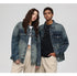 A woman and man are standing close together against a gray backdrop. Both wearing faded blue denim trucker jackets over cropped tops with matching beige pants