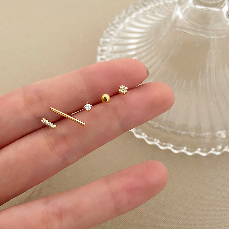 A hand holding a set of six mismatched gold stud earrings, including a bar, ball, square, and cubic zirconia designs, against a neutral background.