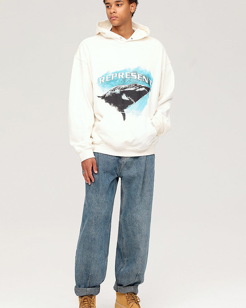 A model wearing white 'REPRESENT' shark graphic hoodie, relaxed-fit blue jeans and tan work boots, posing against white backdrop