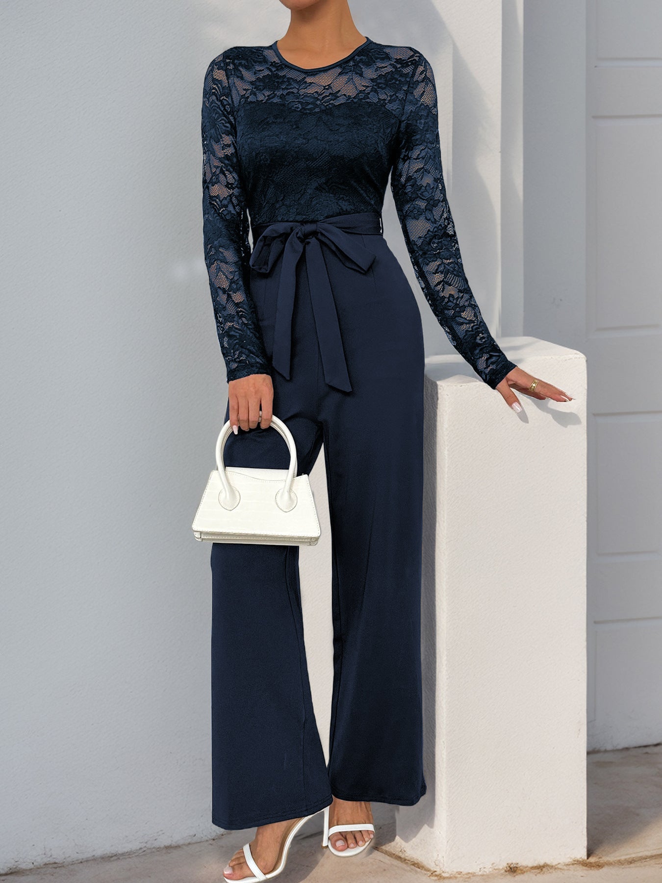 A navy blue version of the lace jumpsuit, styled similarly with white accessories, posing elegantly.
