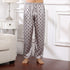 Model wearing silver silk pajama pants with gray polka dot pattern and light gray cuffed hem, standing barefoot on wooden floor next to white nightstand