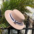 A pink straw boater hat resting on a black metal fence with green palm leaves in the background, featuring a black-and-white ribbon band and silver charm.